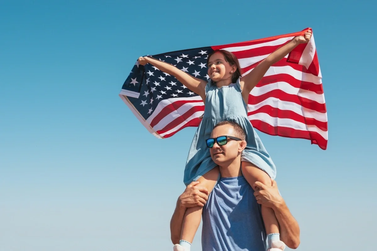 A veteran carries his daughter on his back while she waves an American flag.